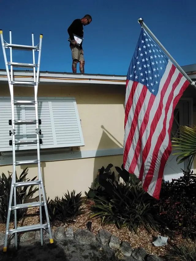 Inspector standing on a ranch-house roof above an aluminium ladder and an American flag