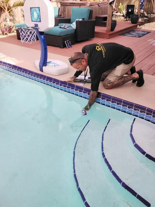 Inspector kneeling at a pool edge checking the waterline tile and deck